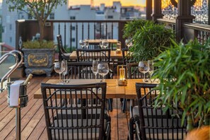 Un área de comedor al aire libre con mesas de madera y sillas de mimbre negro, rodeada de follaje y una barandilla decorativa.