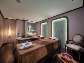 A spa room with two massage tables, a chair, and a small table with rose petals. - Tonkin Luxury Boutique Hotel & Spa (Hanoi)