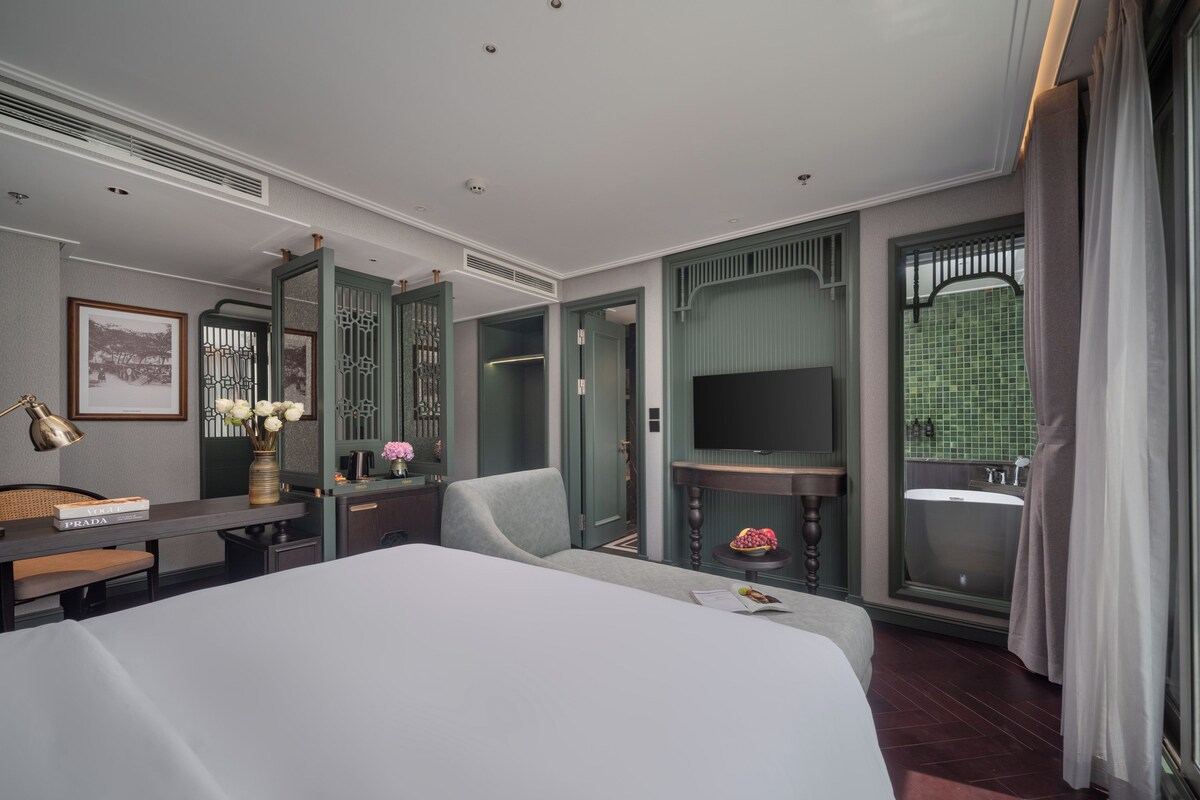 Una habitación de hotel moderna con una cama grande, un escritorio con lámpara, un televisor y un baño con pared de azulejos verdes y una bañera.