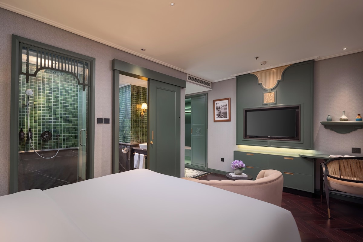 A modern hotel room with a large bed, a desk with a flat-screen TV, a seating area, and a bathroom with a green-tiled shower area.