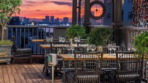 Un área de comedor al aire libre con mesas y sillas, vista al horizonte de la ciudad y decoración en la pared.