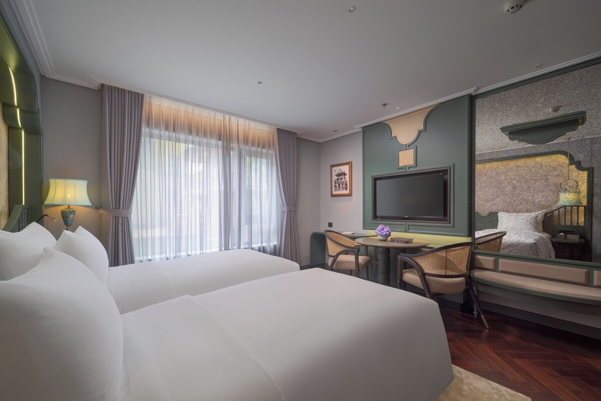 A hotel room with two beds, a TV, a desk with chairs, and a large window with curtains.