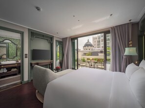 Een hotelkamer met een groot bed, een televisie, een tafeltje met een vaas en een balkon met uitzicht op gebouwen.