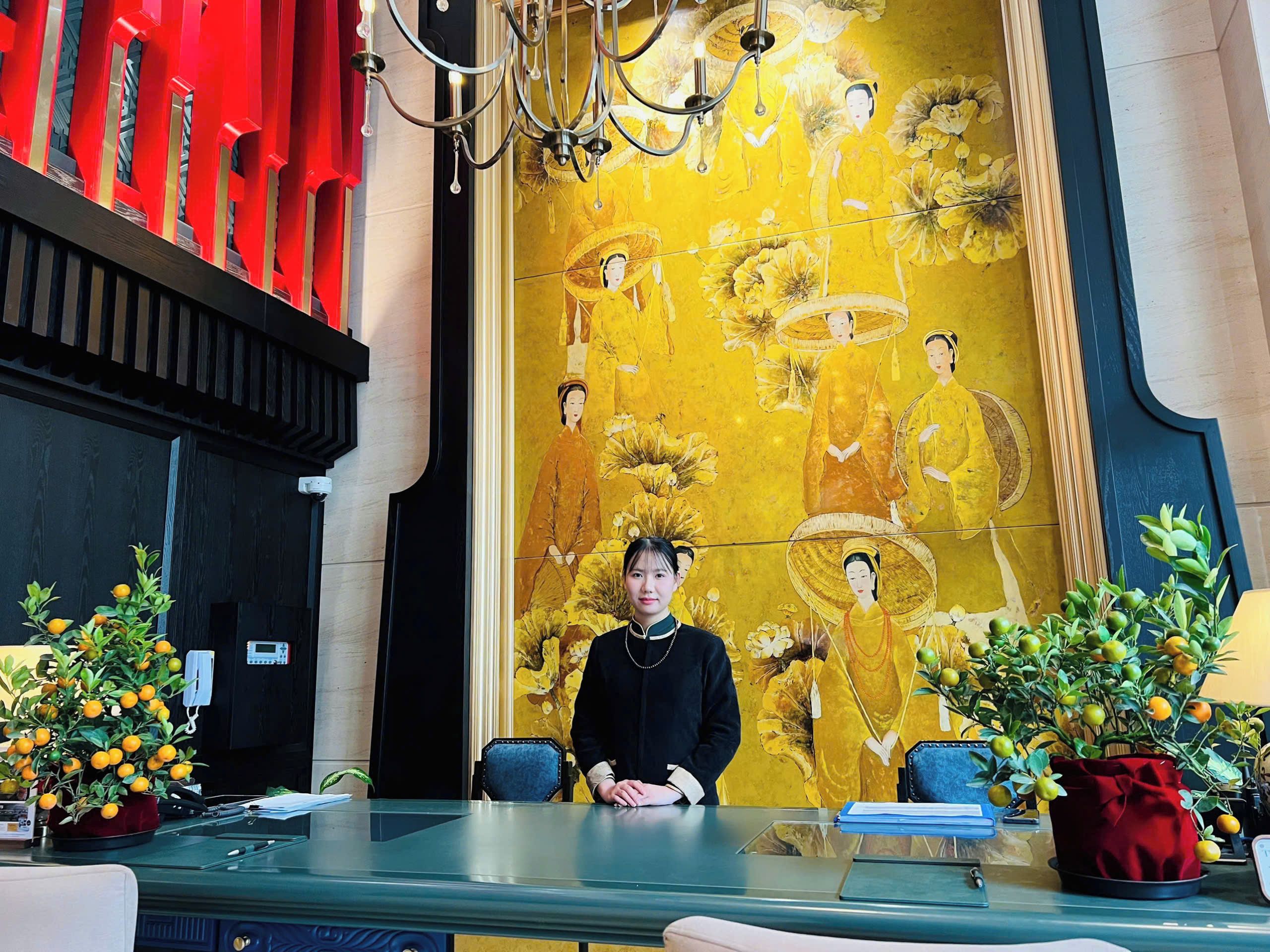 A reception desk with a large mural of traditional figures, a chandelier, and potted plants.