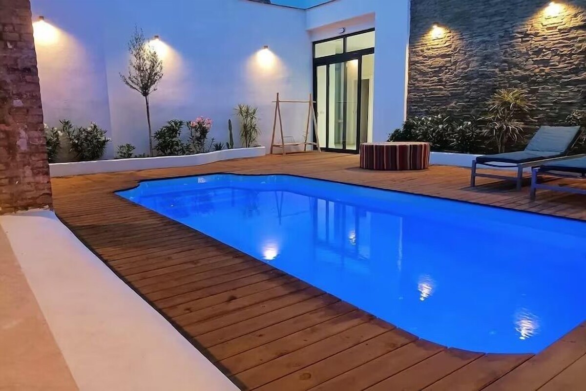 A swimming pool with wooden decking, surrounded by plants and a stone wall.
