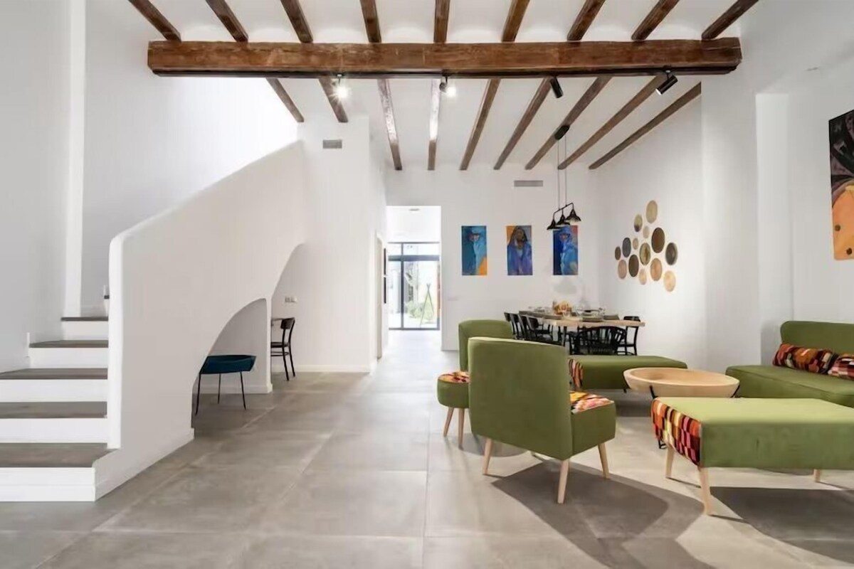 A spacious living area with a staircase, green sofas, a dining table, and artwork on the walls.