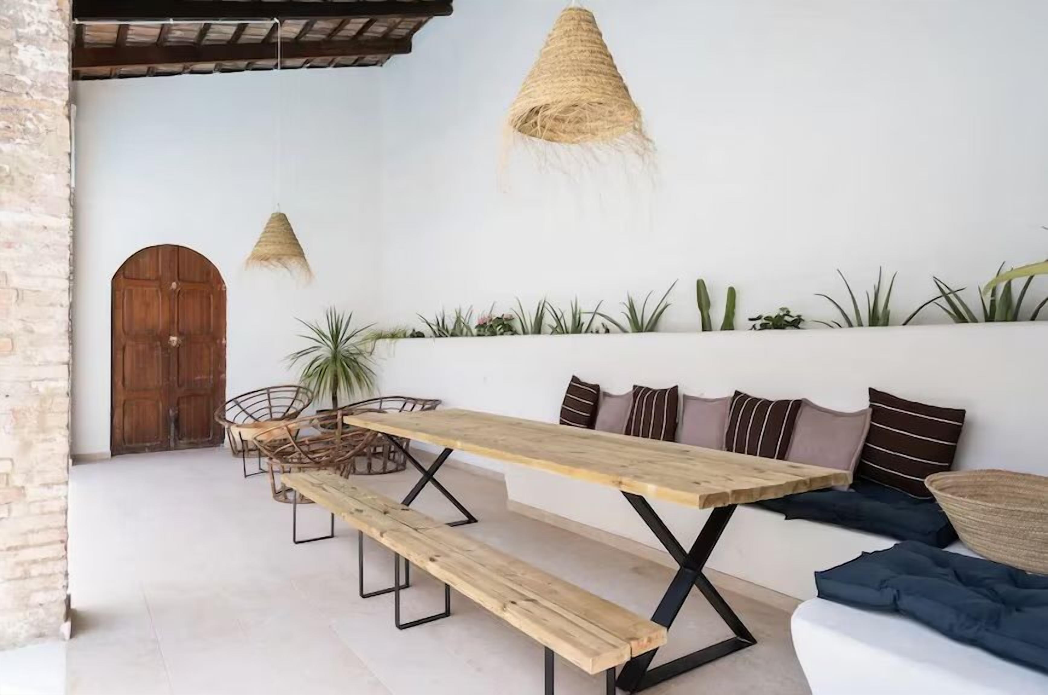 Un espacio al aire libre rústico con una mesa y bancos de madera, una lámpara colgante tejida, y diversas plantas a lo largo de la pared.