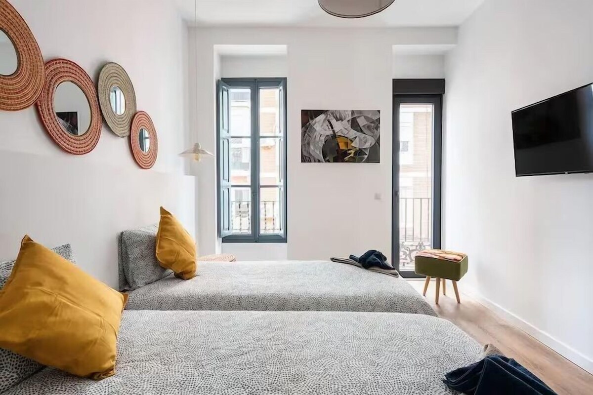 A modern bedroom with two beds, a flat-screen TV, and a large window with a view of buildings.