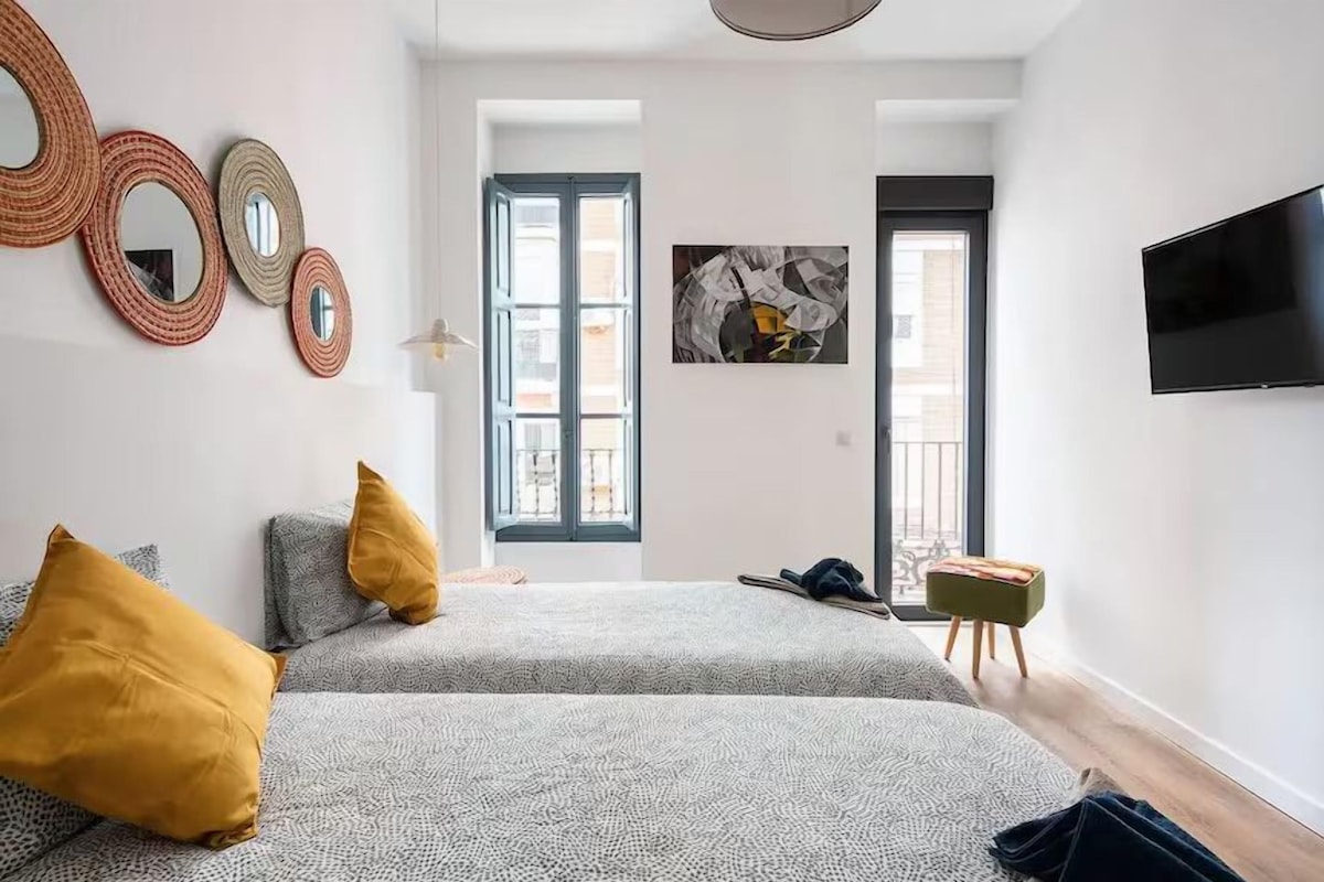 A modern bedroom with two beds, a flat-screen TV, and a large window with a view of buildings.