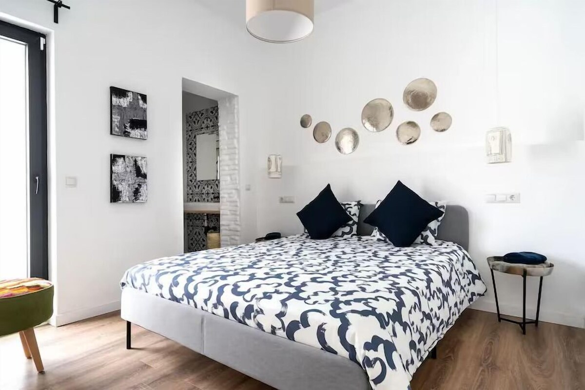 A modern bedroom with a large bed, a small side table, and a view of a bathroom through an open door.