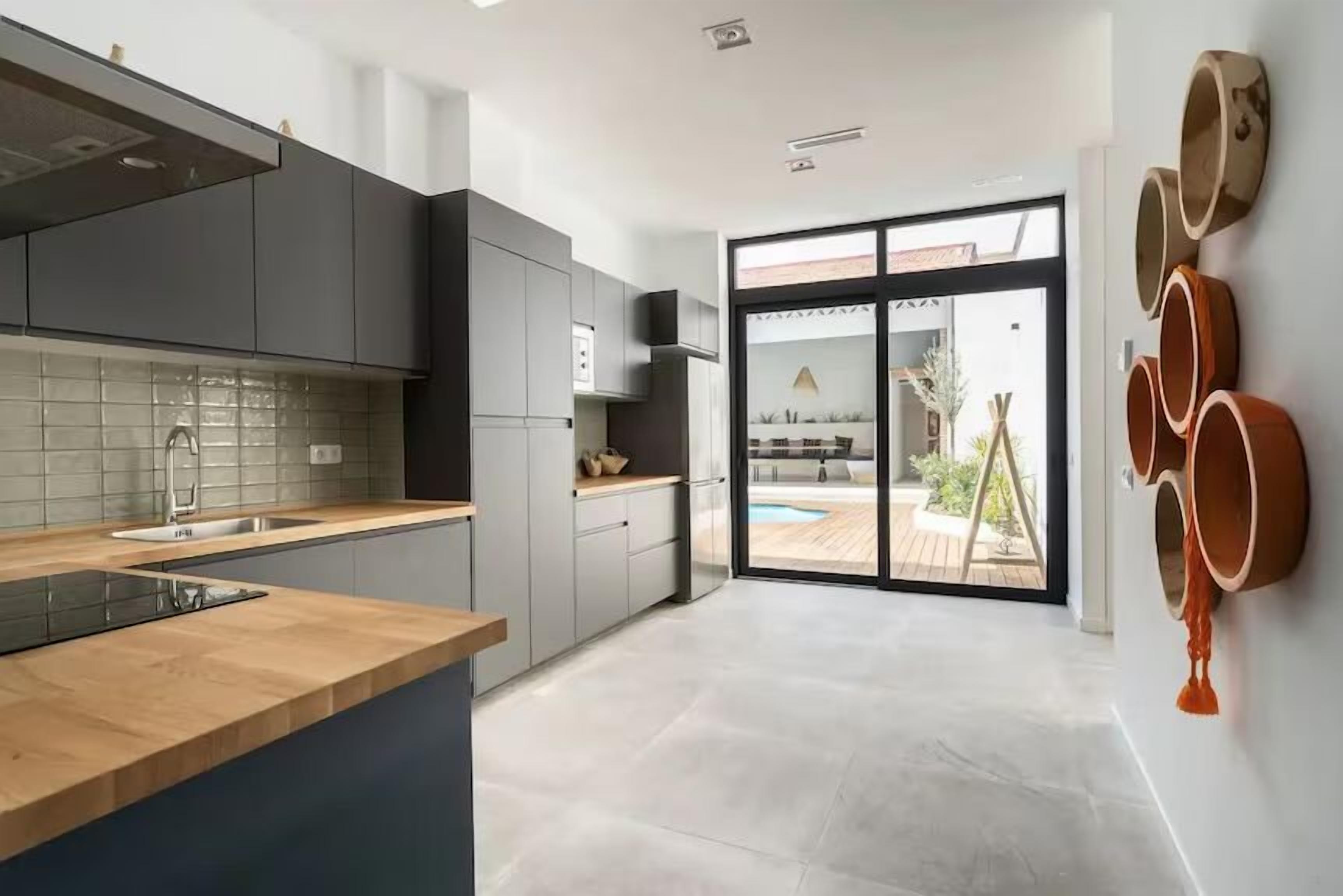A modern kitchen with grey cabinets, a wooden countertop, and a large window overlooking a pool area.