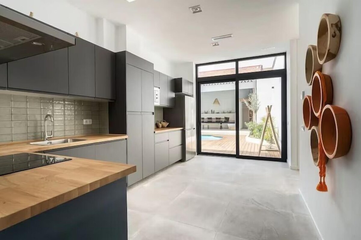 A modern kitchen with grey cabinets, a wooden countertop, and a large window overlooking a pool area.
