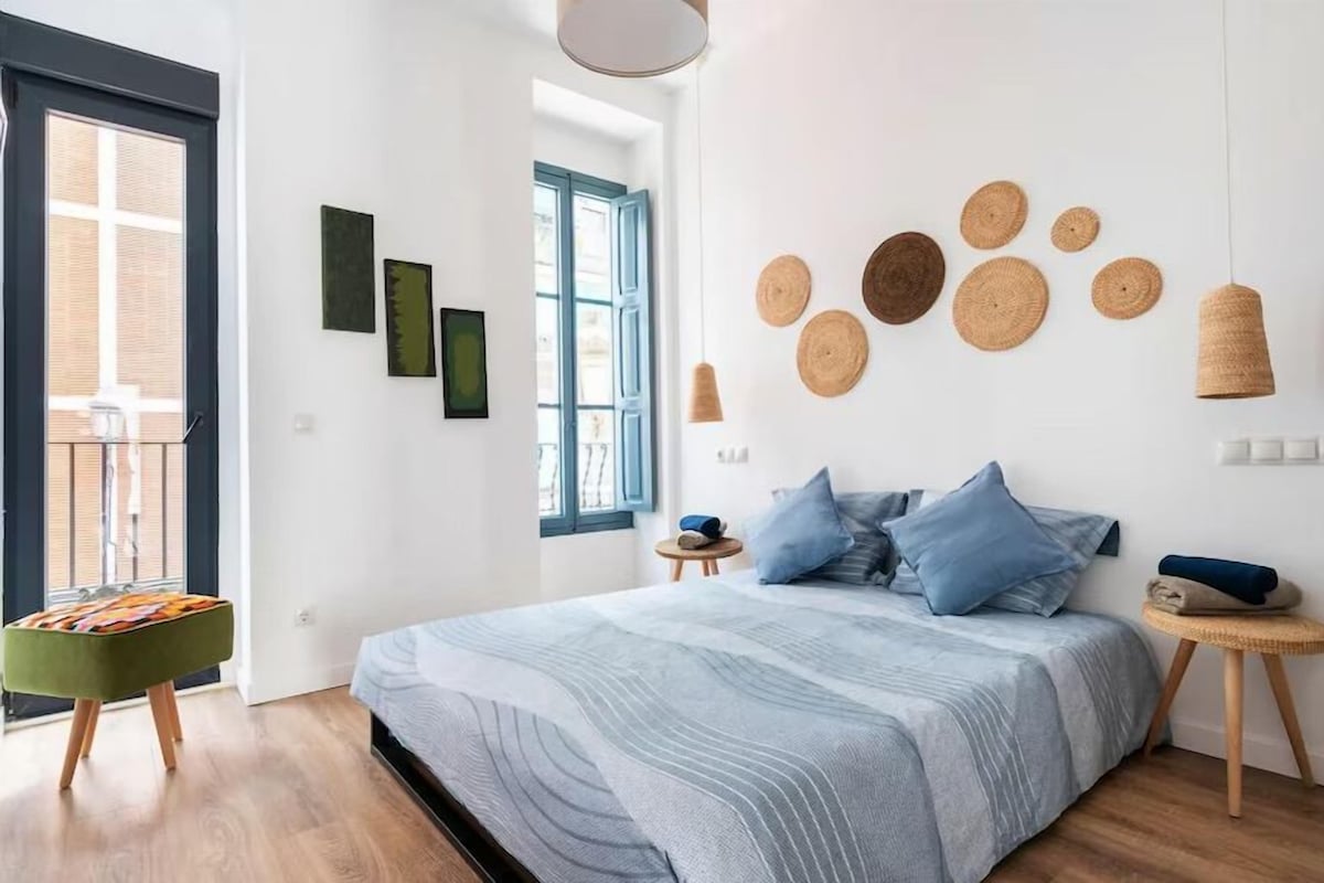 A bedroom with a bed, a small stool, a window, and wall art.