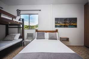 A hotel room with a bunk bed, a single bed, a chair, a nightstand, a painting, and a window with a cityscape view. - Finca Hotel Zona Franca (El Carmen de Viboral)