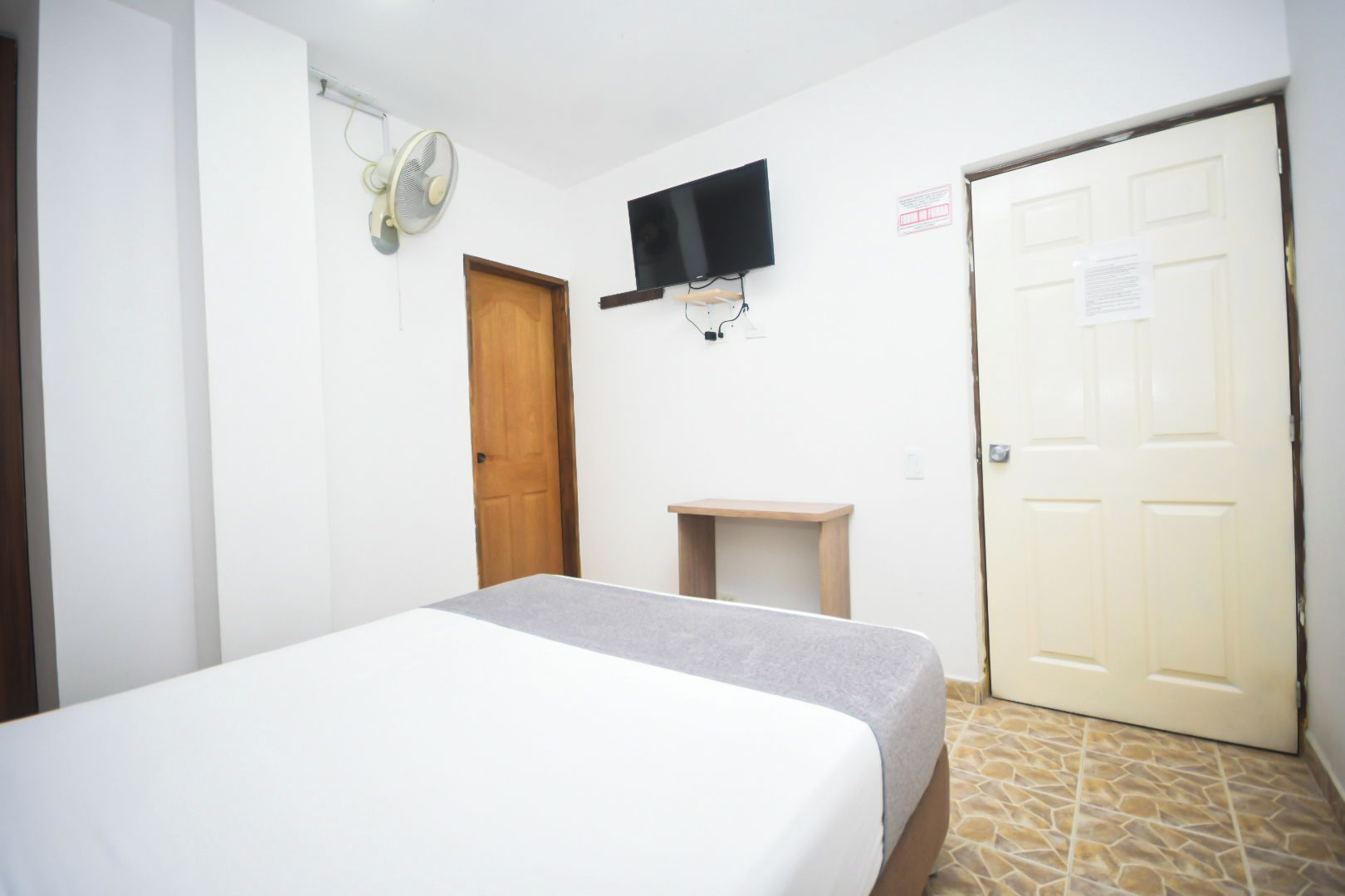 A bedroom with a bed, a wooden door, a small table, a wall-mounted TV, and a fan.
