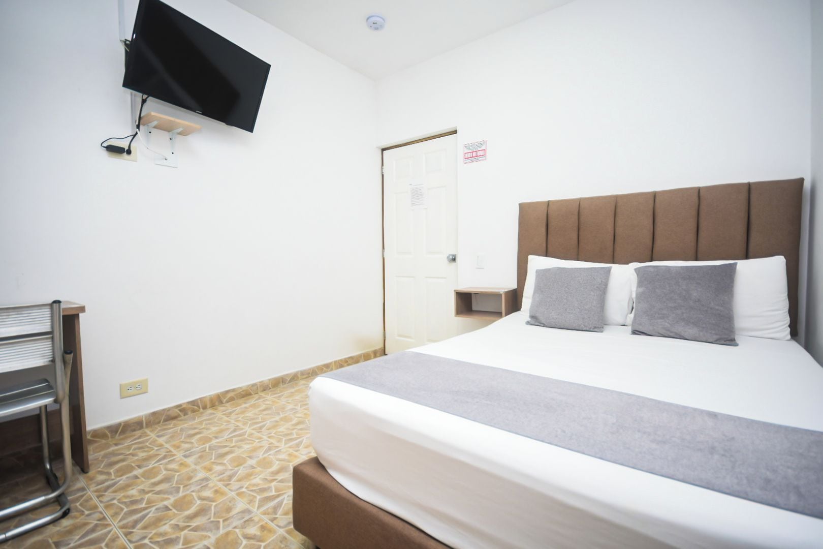 A hotel room with a bed, a TV mounted on the wall, a door, and a small wooden nightstand.