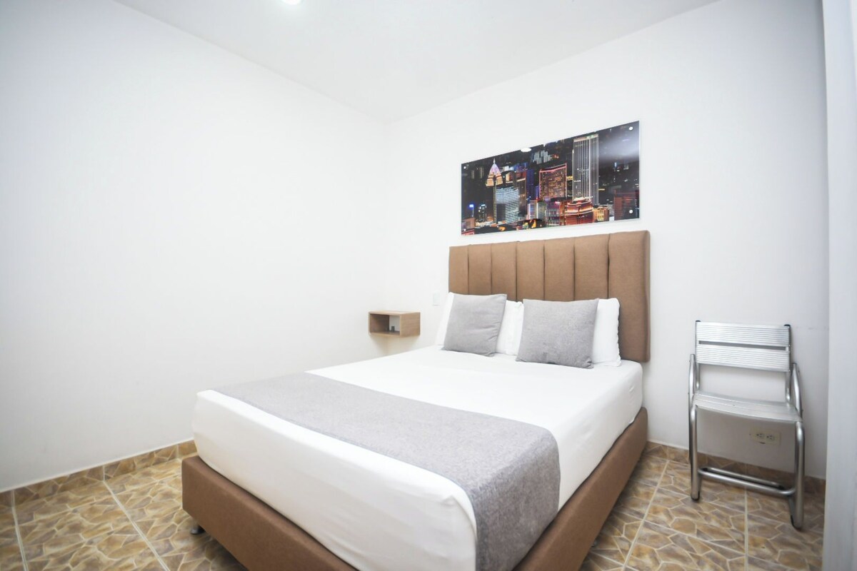Ein Hotelzimmer mit Bett, Nachttisch, Wandlampe und einem Stadtbild.