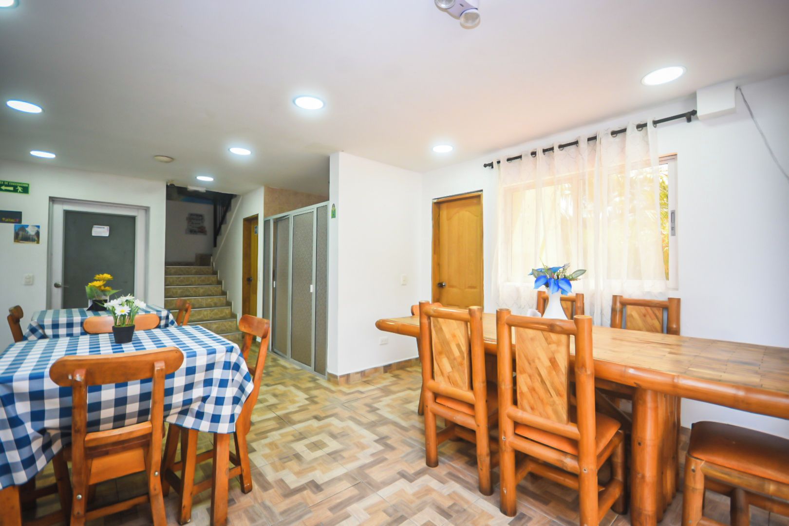 A dining area with a checkered tablecloth, wooden chairs, a vase with flowers, and a staircase in the background.