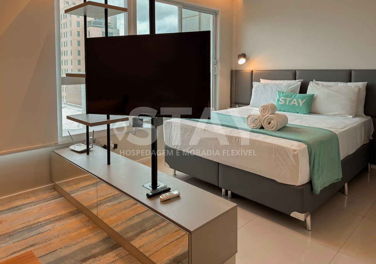 A modern hotel room with a bed, a TV, a desk, and a view of buildings outside.