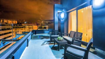 A balcony with wicker furniture and a table, overlooking a cityscape at night.