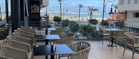 Uma área externa com assentos de vime e mesas, com vista para uma praia com palmeiras e céu claro.