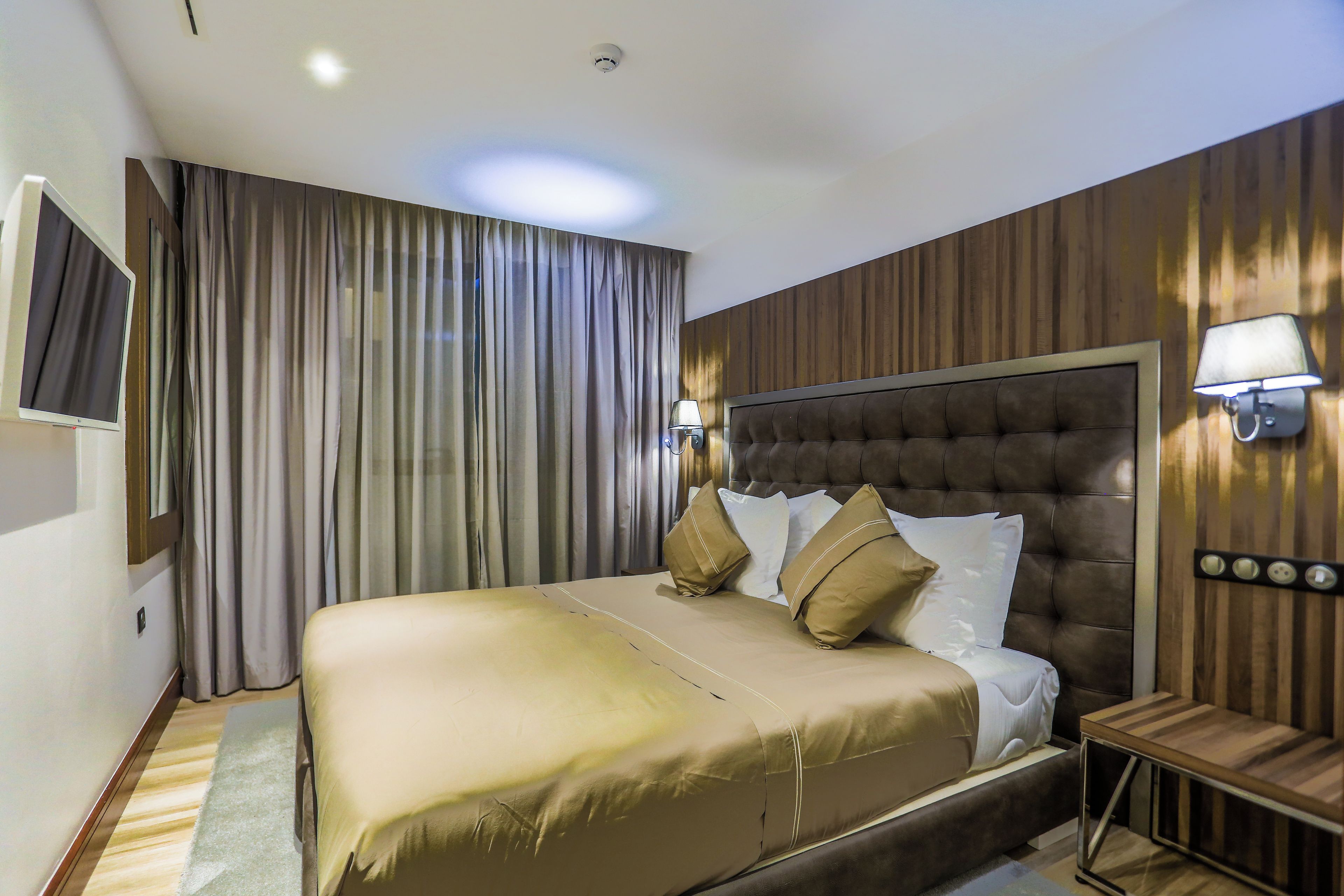 A hotel room with a large bed, a flat-screen TV, a wooden headboard, and a nightstand with a lamp.