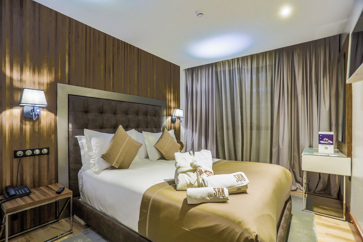 A hotel room with a large bed, a bedside table with a lamp, a nightstand with a phone, and a wall-mounted clock.