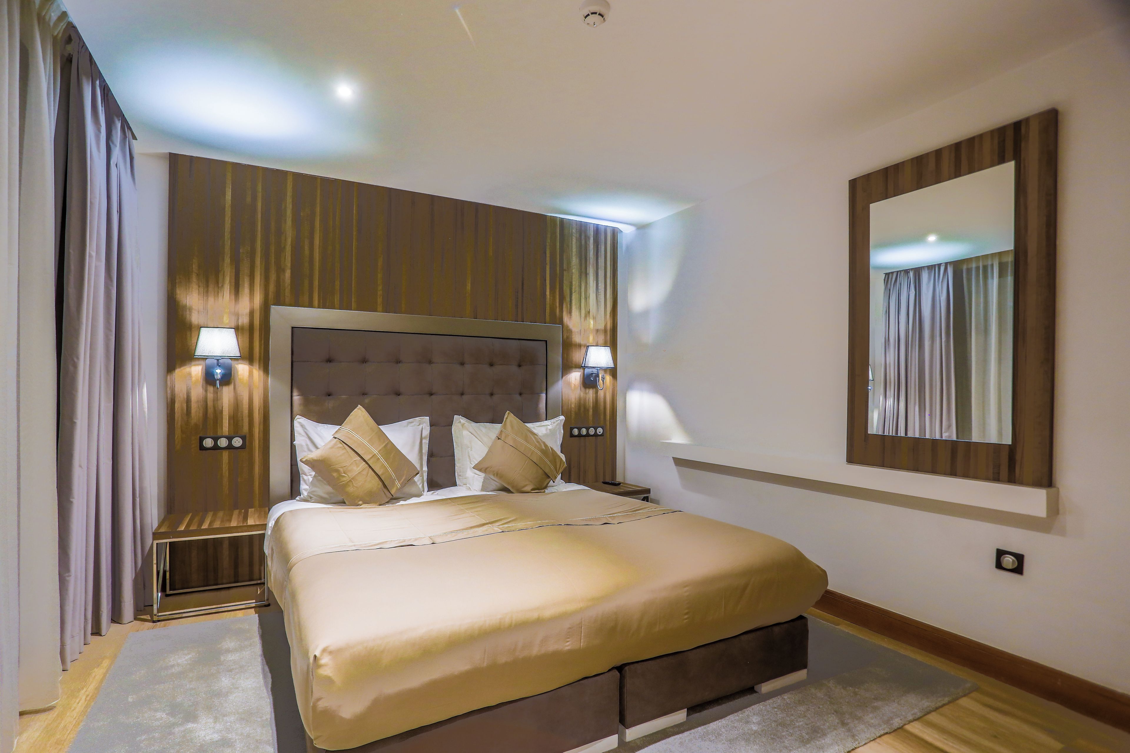 A hotel room with a large bed, two bedside lamps, a headboard, a mirror, and curtains.