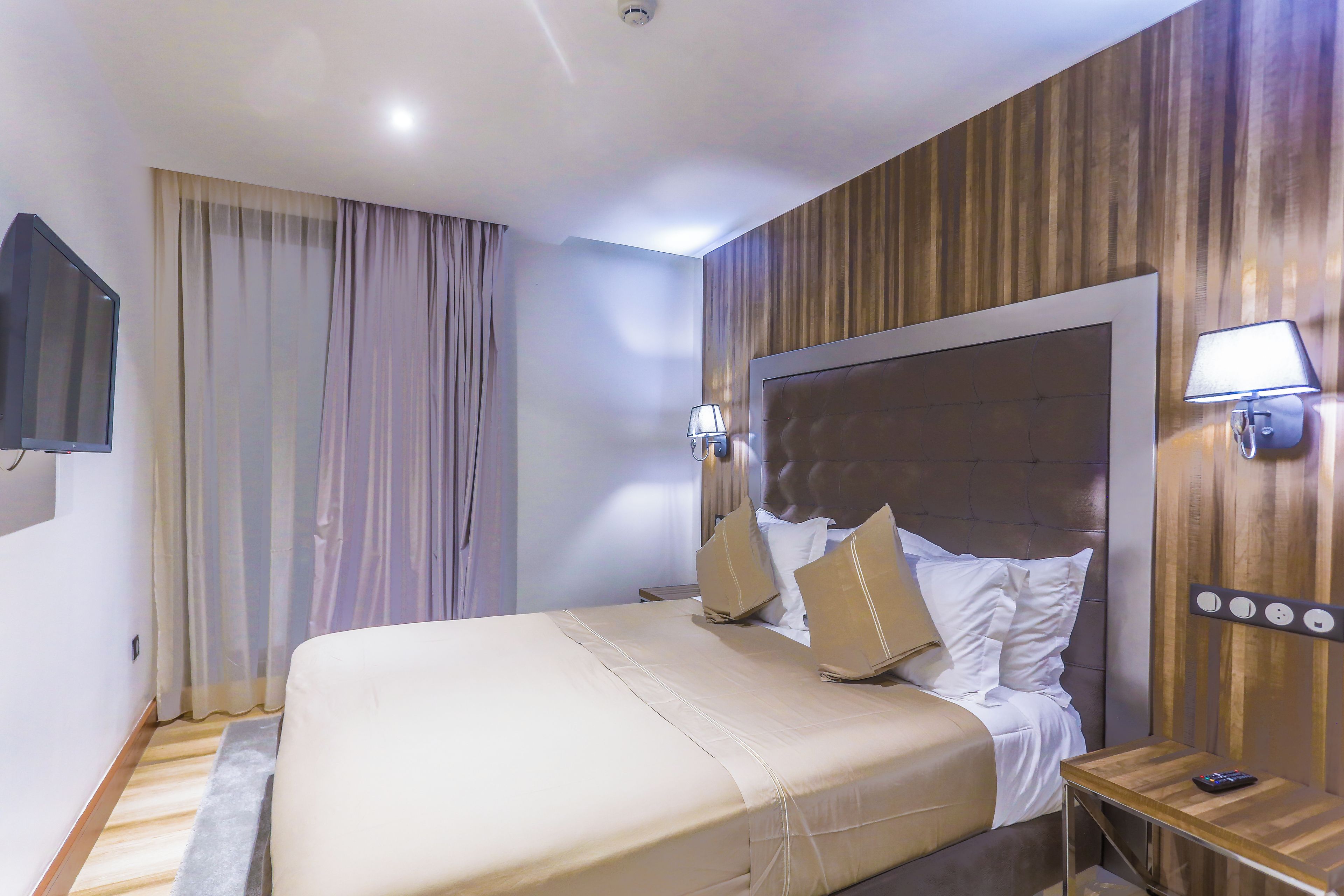 A hotel room with a large bed, wooden paneling on the walls, a mounted TV, and a bedside table with a lamp.