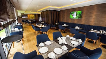 A restaurant with tables set for dining, blue chairs, and a TV on the wall.