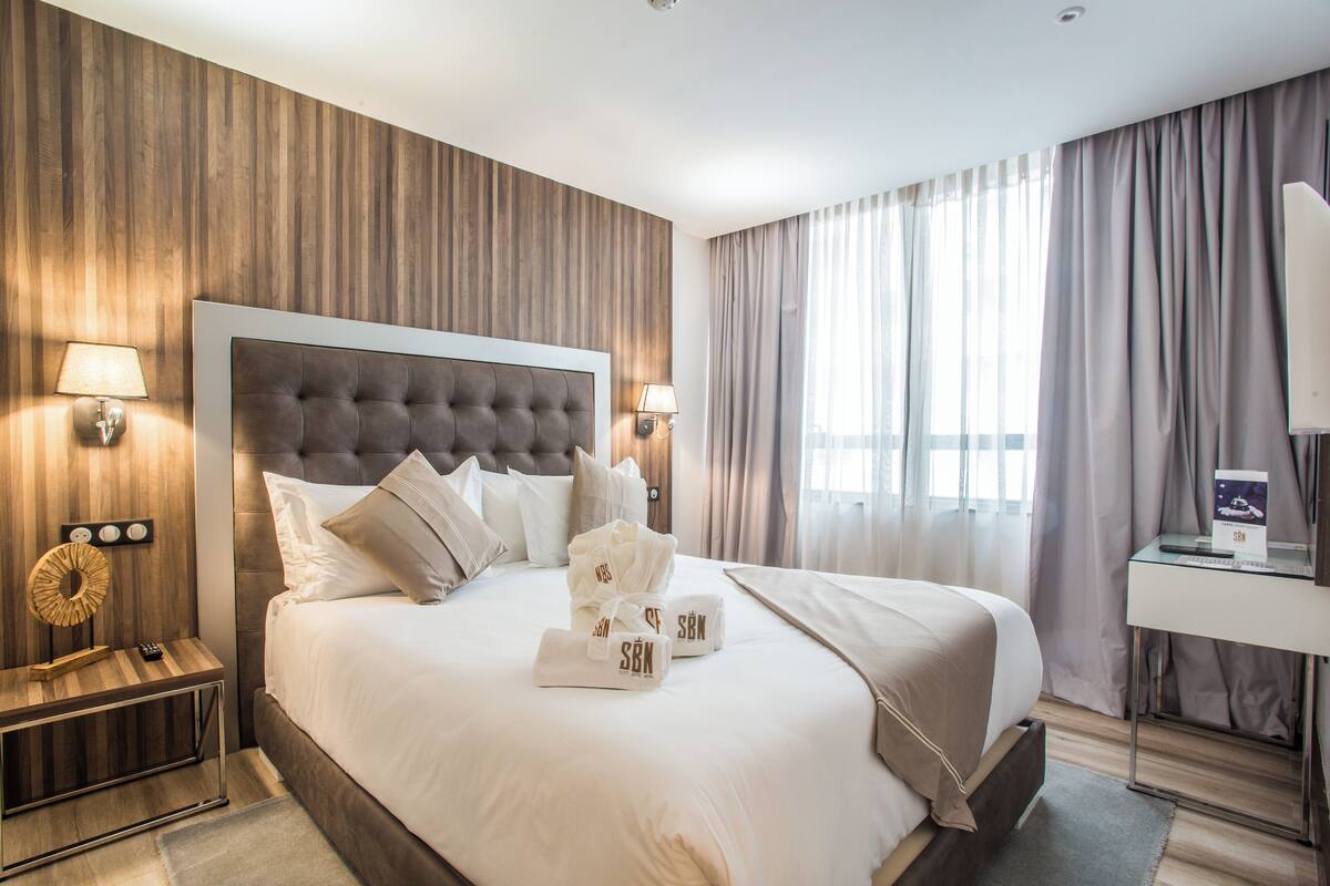 A modern hotel room with a large bed, wooden panel walls, and a view of the city through large windows.