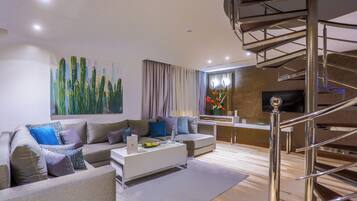 A modern living room with a sofa, coffee table, and a TV. There is a staircase with a metal railing leading to an upper level.