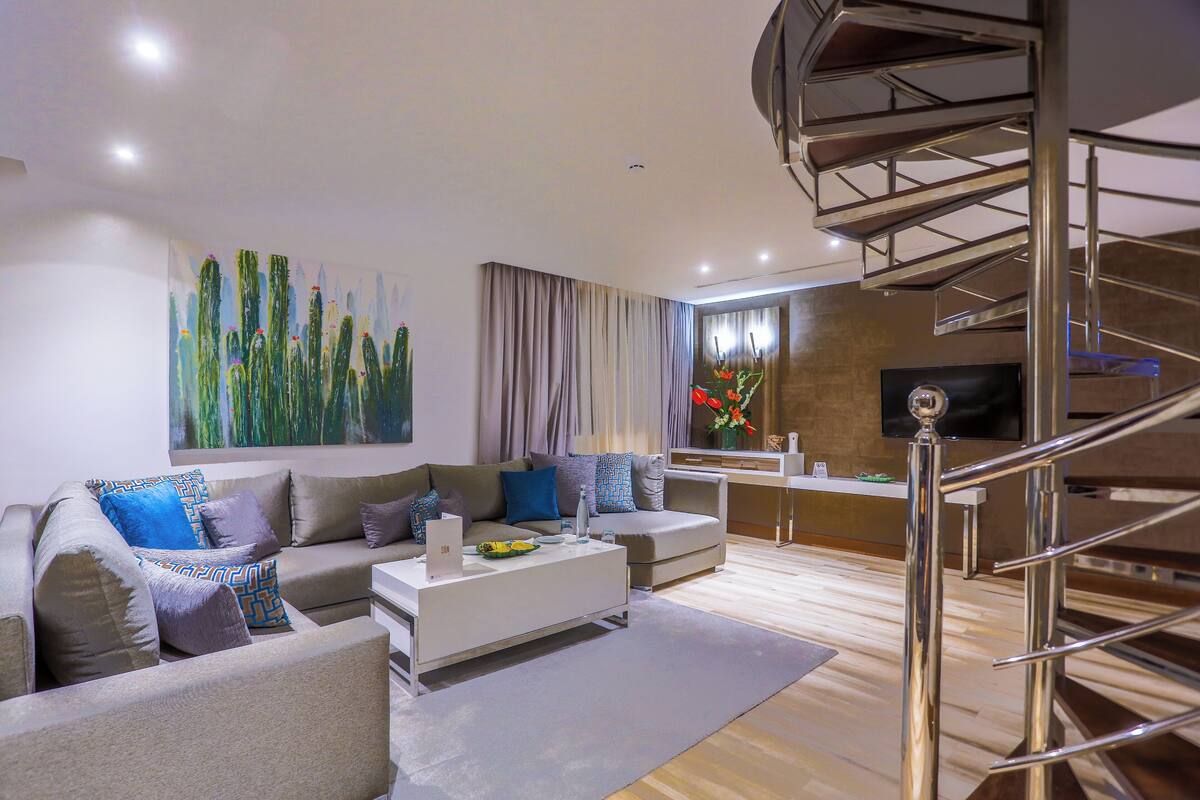 A modern living room with a sofa, coffee table, and a TV. There is a staircase with a metal railing leading to an upper level.