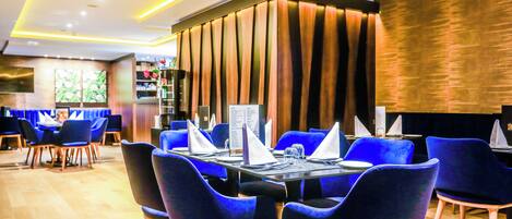 Een restaurant met een modern interieur, met blauwe beklede stoelen, houten tafels en een decoratief wandpaneel.