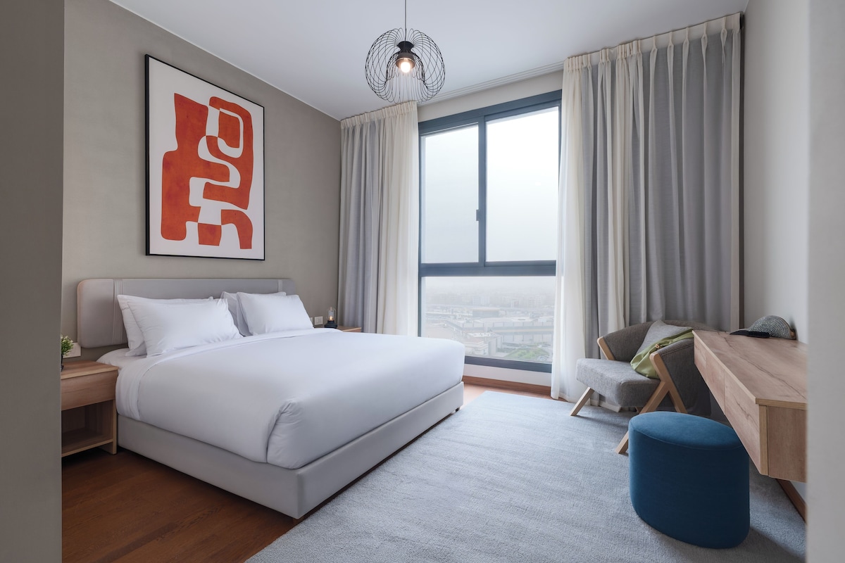 Una habitación de hotel moderna con una cama grande, un escritorio, una silla y vistas a la ciudad desde la ventana.