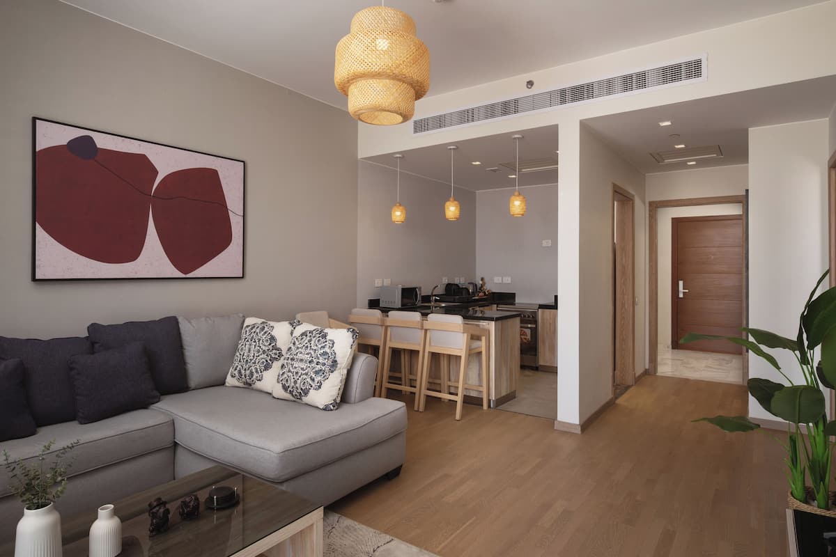 Una sala de estar moderna con un sofá gris, una mesa de centro y un cuadro abstracto grande. Hay una zona de cocina con una barra y lámparas colgantes.