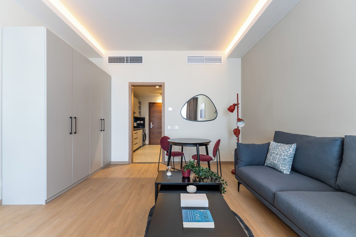 A modern living room with a grey sofa, a round table, and red chairs. There is a large mirror on the wall and a small table with a plant and books.