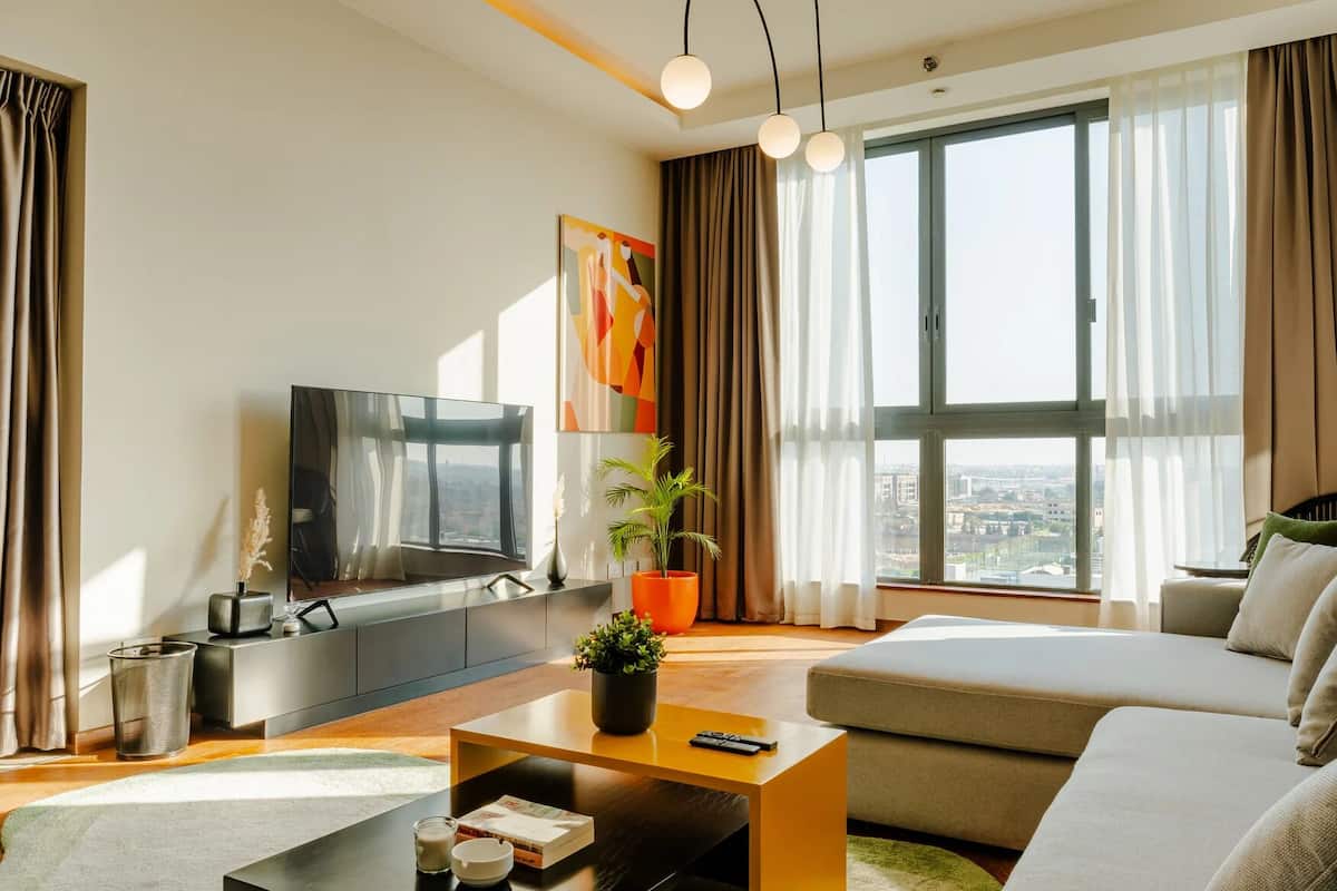 Una sala de estar moderna con un televisor grande, un sofá, una mesa de centro y vistas a la ciudad a través de amplios ventanales.