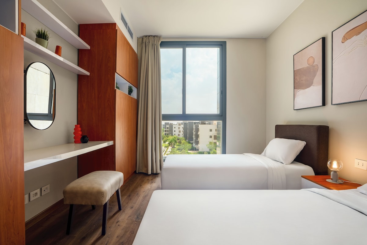 Habitación de hotel con dos camas, un banco de madera, un espejo, un estante con adornos y un amplio ventanal con vista a la ciudad.