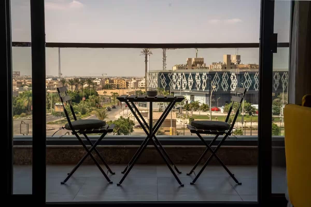 Un balcón con dos sillas y una mesa, con vista a un paisaje urbano que incluye edificios y un puente.