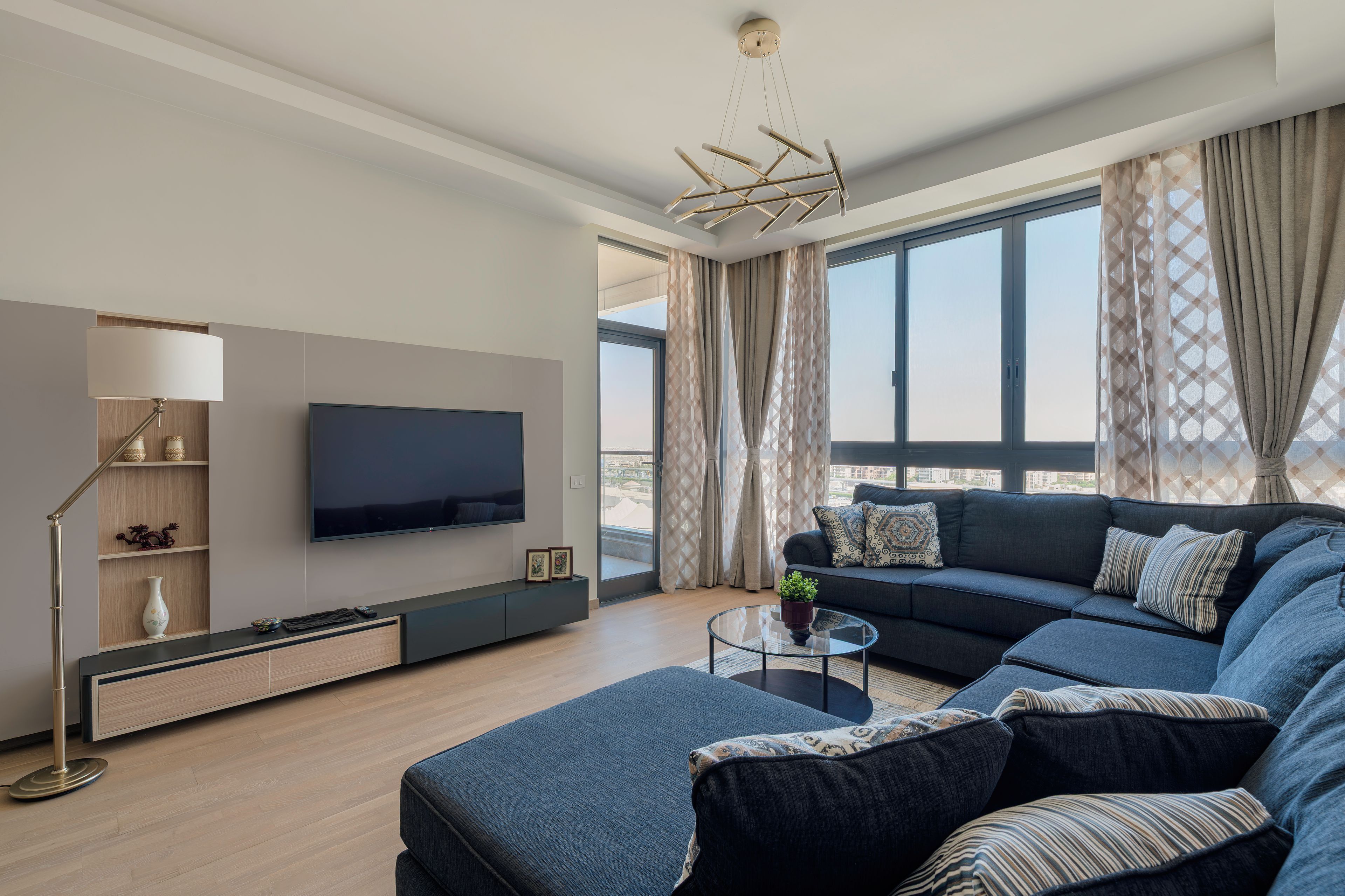Un salon moderne avec un canapé d’angle, un téléviseur à écran plat fixé au mur, une table basse en verre et un lampadaire.