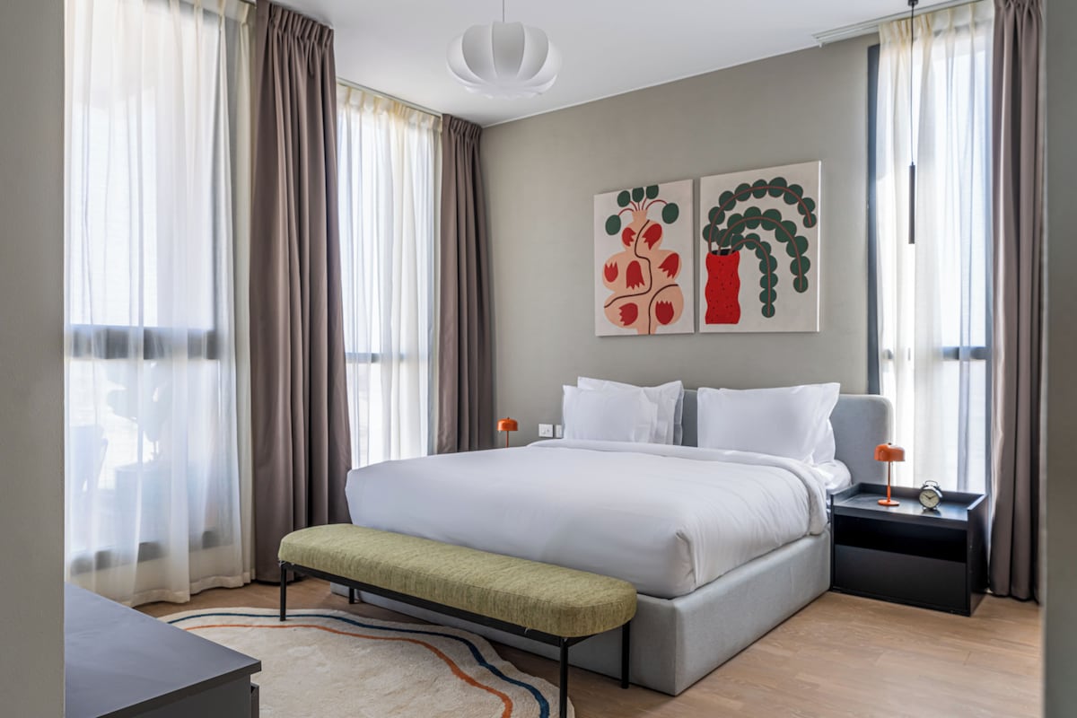 Una habitación de hotel moderna con una cama grande, un banco, un escritorio y una mesita de noche con lámpara y despertador.