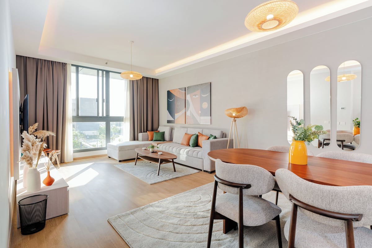 Una sala moderna con un sofá, una mesa de centro, una mesa de comedor y sillas. Hay un ventanal con cortinas, un cuadro en la pared y un espejo.