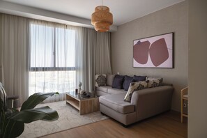 A modern living room with a sofa, coffee table, and a large window with sheer curtains.