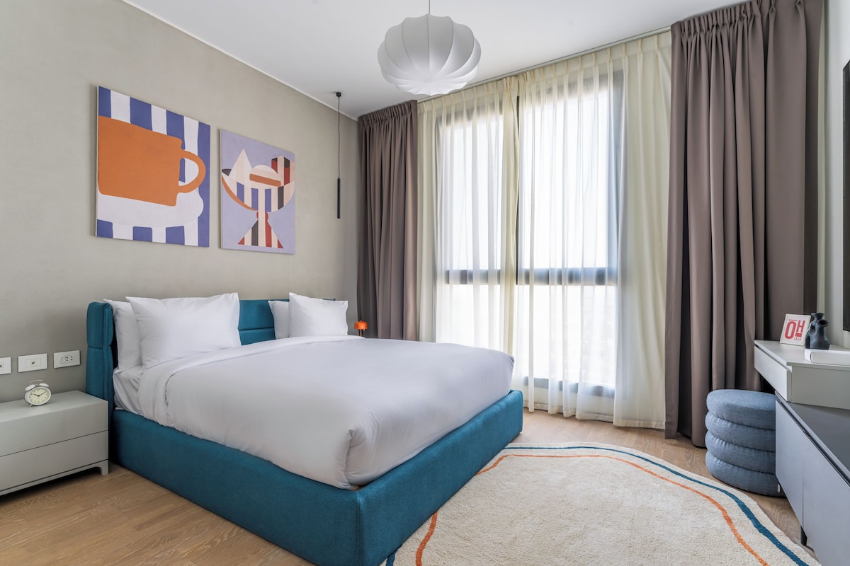 A modern bedroom with a large bed, a nightstand, a desk, and a window with curtains.