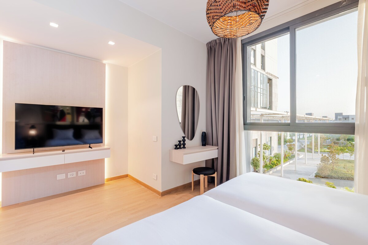 Una habitación de hotel moderna con una cama grande, un televisor montado en una consola blanca, un escritorio pequeño con espejo y vistas a la ciudad a través de amplios ventanales.