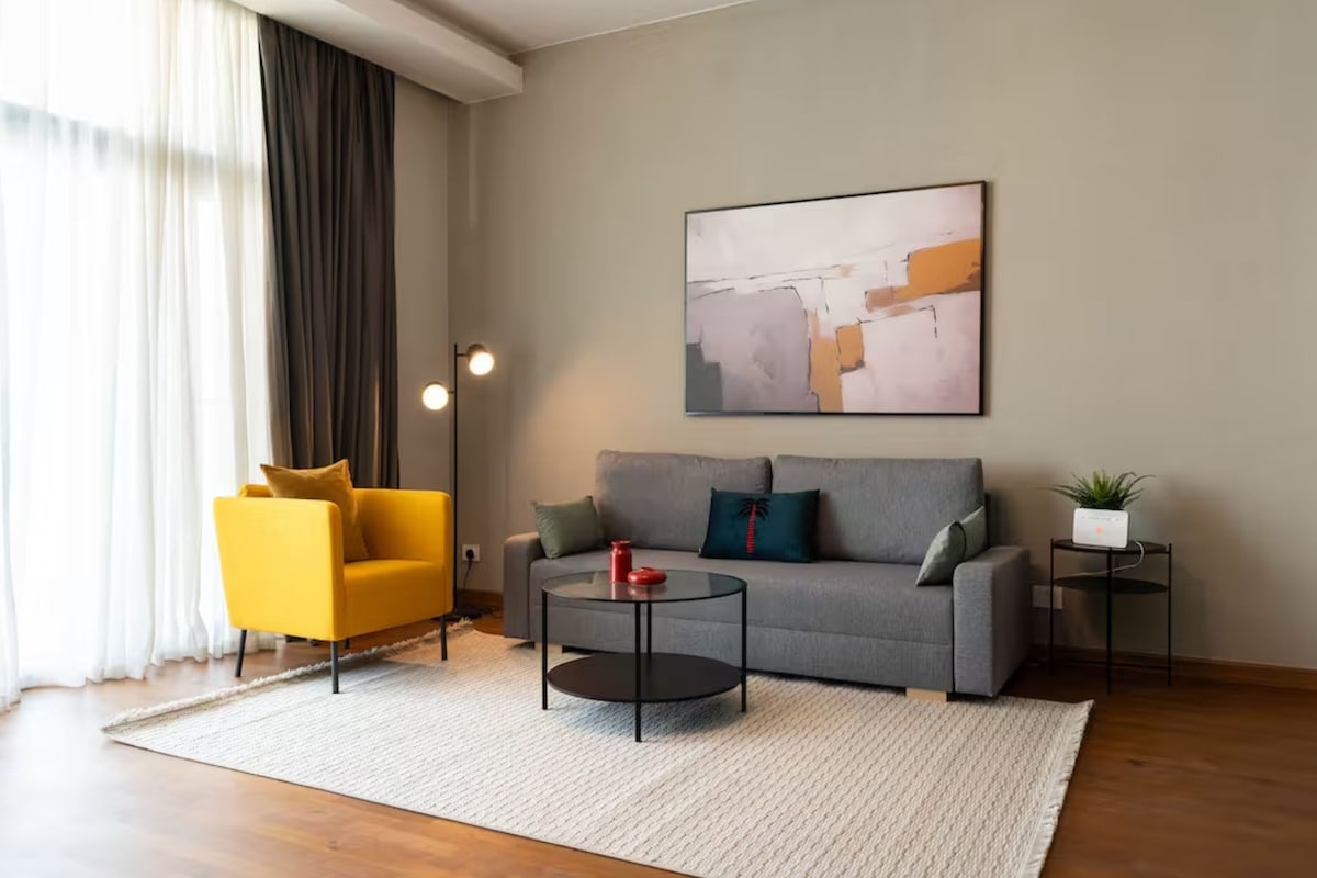 A modern living room with a grey sofa, a yellow armchair, a glass coffee table, and a painting on the wall.