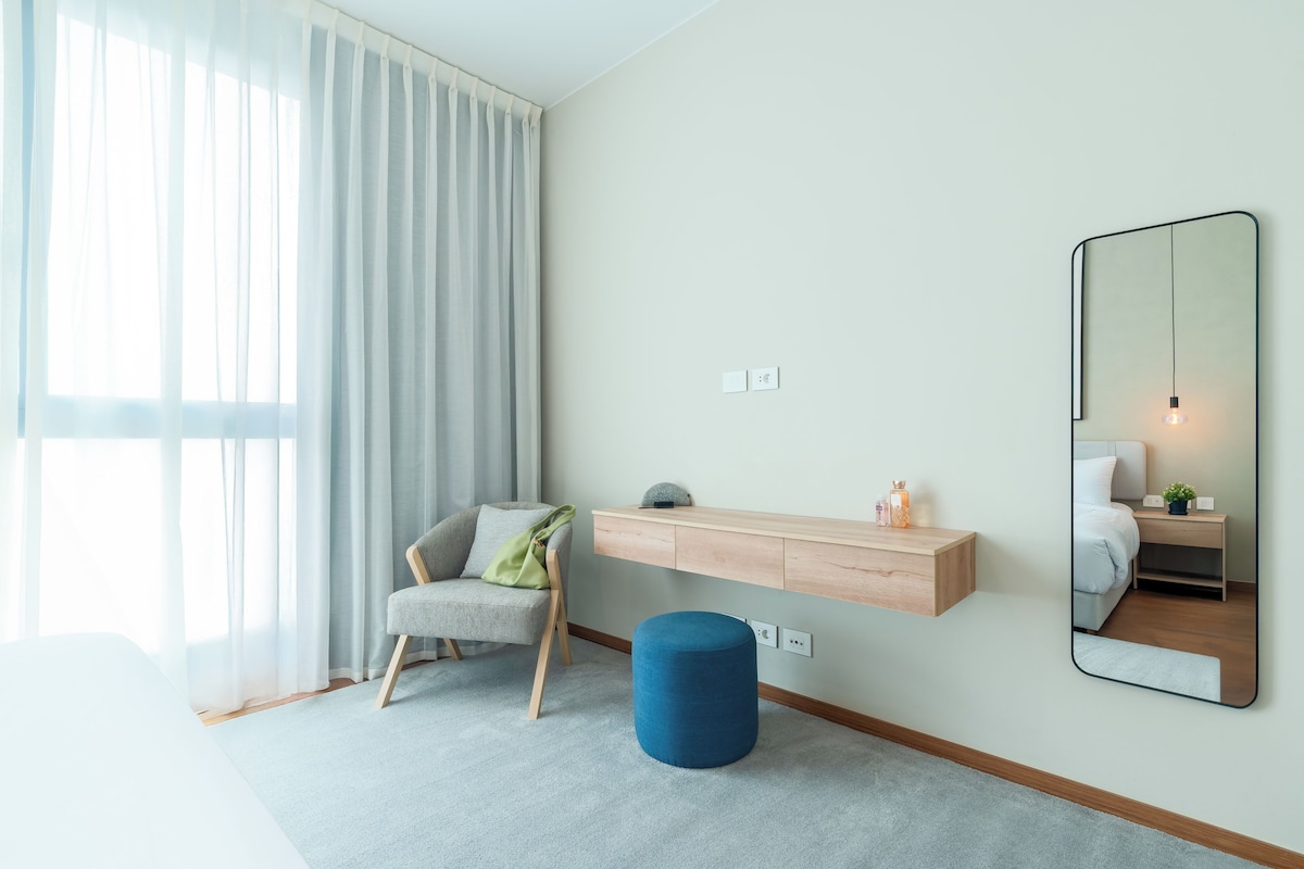 A modern hotel room with a large window, a chair, a bedside table, a mirror, and a blue ottoman.