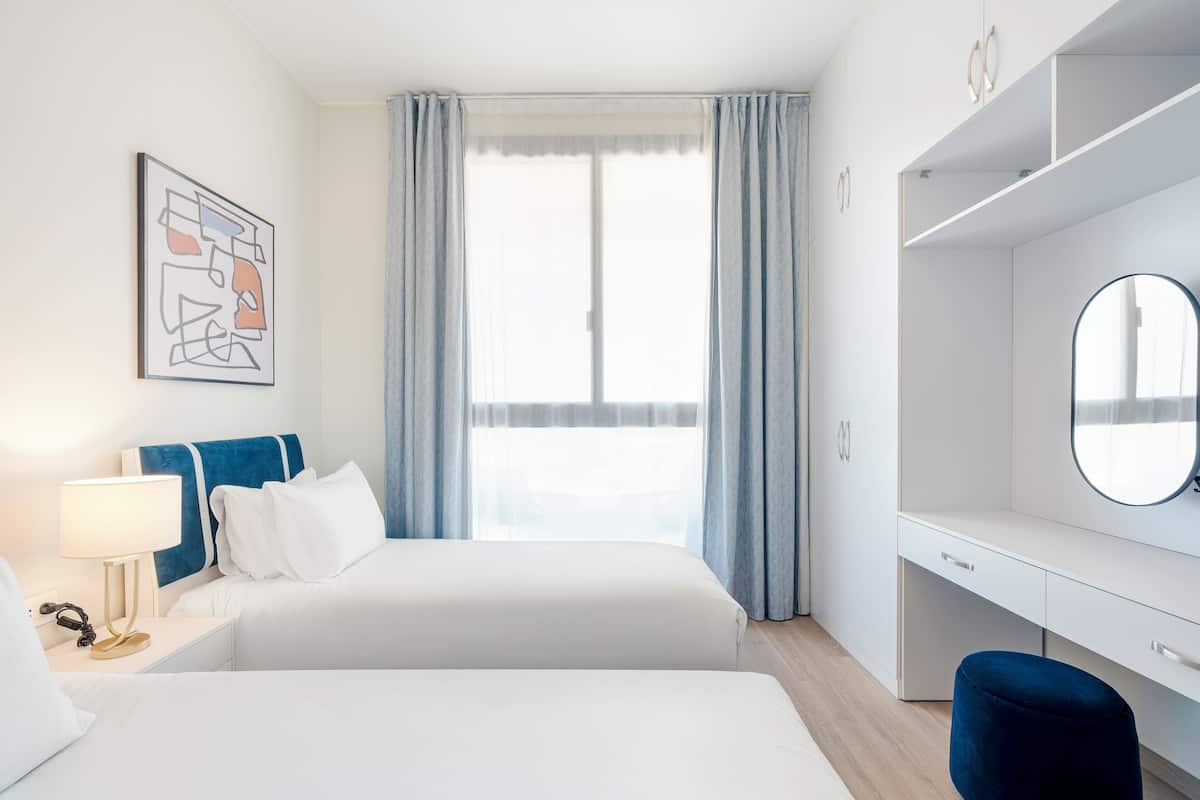 Habitación de hotel con dos camas, un escritorio, una silla, una lámpara, un espejo y una ventana con cortinas.