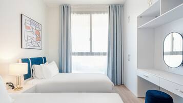 Ein Hotelzimmer mit zwei Betten, einem Schreibtisch, einem Sessel, einer Lampe, einem Spiegel und einem Fenster mit Vorhängen.